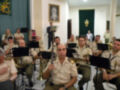 Un grupo de músicos militares toca instrumentos en una sala con decoración formal. En el fondo, se puede ver un retrato de una figura religiosa y una bandera nacional. Los músicos están sentados en sillas, con instrumentos de viento y percusión.
