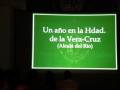 Presentación en un auditorio con una proyección de texto verde sobre fondo negro. Texto: "Un año en la Hdad. de la Vera-Cruz (Alcalá del Río)". Nombre: "Andrés Martínez Quiles".