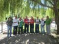 Un grupo de siete personas, seis hombres y una mujer, posan juntos en un parque con árboles verdes. Todos llevan camisetas de fútbol y pantalones, excepto la mujer que lleva una camisa verde. Algunos sostienen trofeos de fútbol, lo que sugiere que han ganado un torneo. La escena está iluminada por el sol, y hay una valla de madera en el fondo.