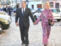 Imagen muestra a una pareja caminando por una calle con edificios y coches al fondo. La mujer lleva un vestido floral y la chaqueta del hombre es de traje negro.