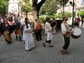 Personas vestidas con trajes tradicionales caminan por una calle, mientras otros observan. La escena parece ser parte de un evento cultural o festivo en una ciudad con edificios históricos al fondo.