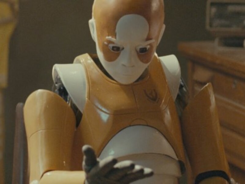 Un robot con traje amarillo y blanco, sentado en una silla, en un entorno que parece ser un laboratorio o taller.