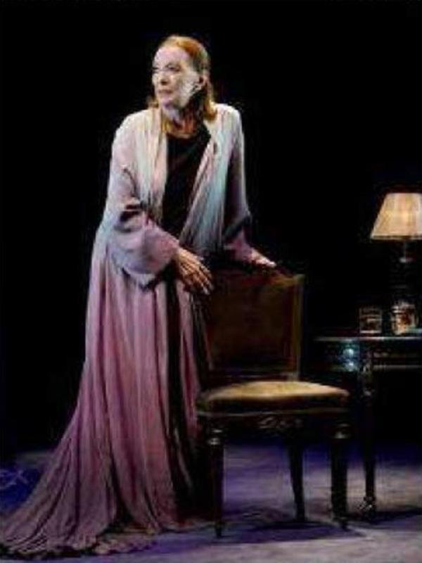 Actriz en escena con vestimenta dramática, posando junto a una silla y una lámpara de mesa.
