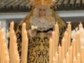 Una figura vestida con un manto dorado y negro, rodeada de velas encendidas. La figura lleva un halo dorado y una corona, lo que sugiere un personaje importante o religioso. La escena parece ser parte de una celebración o procesión, dada la presencia de velas y el atuendo elaborado.