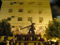 Una imagen nocturna de una procesión religiosa con un paso central que sostiene la cruz, rodeado por fieles y luces. El fondo es un edificio con ventanas y balcones, iluminado por faroles y luces.