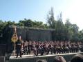 Una ceremonia militar en un escenario al aire libre con un hombre en uniforme de frente, seguido por una fila de soldados sentados. El escenario está decorado con un fondo negro y un estandarte dorado en la izquierda. El cielo es claro y el ambiente parece ser un día soleado.