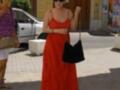 Una mujer en un vestido rojo largo y sandalias de tacón, caminando por una calle con un bolso negro en la mano. Detrás de ella, hay un coche blanco y una tienda con una puerta de madera.