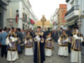 Processión religiosa en una calle de la ciudad, con participantes vestidos con trajes tradicionales y guantes blancos.