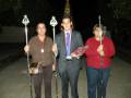 Tres personas con bastones de procesión, una mujer y dos hombres, posan en la noche. La imagen muestra detalles como sus ropa formal y las insignias de procesión que llevan en los bastones.