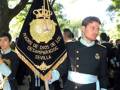 Un soldado de uniforme negro y azul sostiene una bandera con el escudo de la Real Armada de España y el nombre 'Madre de Dios de los Desamparados' en Sevilla.