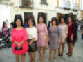 Una fila de mujeres vestidas con diferentes estilos de falda y blusa, posando frente a una casa blanca con balcones.