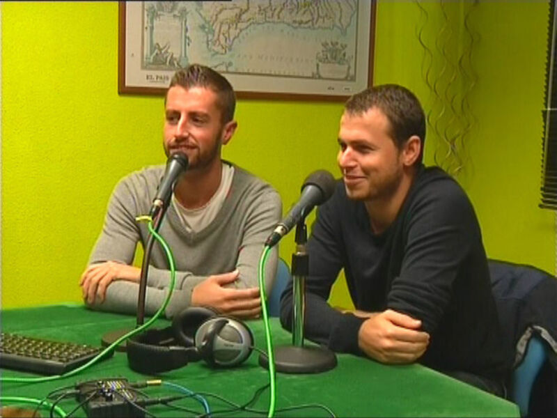 Dos hombres sentados en una mesa con micrófonos y auriculares, posiblemente en un estudio de radio o podcast.