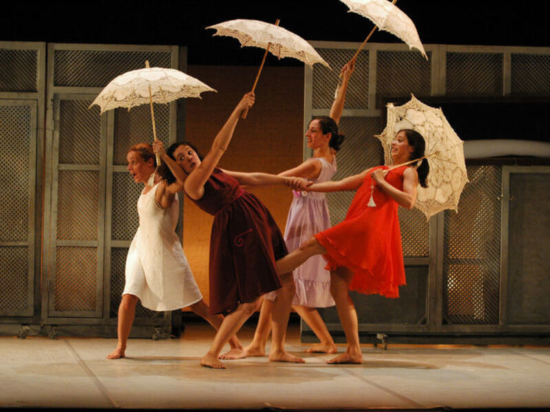 Cuatro bailarinas en escenario, vestidas con vestidos de colores y sosteniendo paraguas blancos, realizando una coreografía sincronizada.