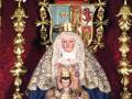 Imagen de una Virgen con el Niño Jesús, vestida con trajes tradicionales y adornada con un manto bordado, en un altar decorado con elementos dorados y un escudo realista.