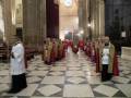 Una procesión religiosa en una catedral con sacerdotes vestidos de rojo y blanco, caminando en fila. El suelo es de azulejos blancos y negros, con columnas y ventanas en el fondo.