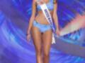 Una modelo en traje de baño azul con un sash que dice "Miss Barbados" camina por el escenario de Miss Universo. El fondo es un diseño abstracto con colores azul y blanco.