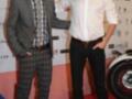 Dos hombres posan en un evento, uno con traje y corbata, el otro con camisa blanca y pantalones. El fondo muestra logos de empresas.