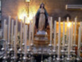 Una estatua de la Virgen María se encuentra en un altar rodeada por velas encendidas. El ambiente es reverente y decorado con detalles de lujo, incluyendo un aire acondicionado en la parte superior derecha.