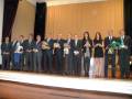 Una fila de personas en trajes formales, posando para una foto frente a cortinas doradas. Algunos sostienen flores y medallas, indicando un evento de reconocimiento o premiación.