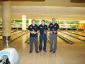 Tres hombres celebran en una alineación de bowling con trofeos en mano, posando juntos en una alineación de bowling.