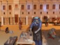 Un diorama que muestra una escena histórica con un edificio colonial en el fondo. En primer plano, varias figuras antropomórficas realizan diferentes actividades en torno a una mesa. La escena parece representar un momento de la vida cotidiana en un lugar colonial, posiblemente en el siglo XVIII o XIX.