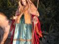 Una figura vestida con una capa roja y un manto azul, posiblemente representando a la Virgen María, sostiene una vara y está de pie junto a un animal. La escena parece ser parte de una representación navideña o religiosa, con un fondo oscuro y árboles en el horizonte.