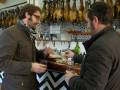 Two men tasting food in a restaurant with hanging cured meats in the background.