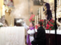 Una imagen religiosa en un altar con una persona bendiciendo a otra, rodeados de flores y velas.