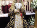 Imagen de una Virgen María vestida en traje tradicional, con un manto dorado y oro, sentada en una silla de plata. La imagen está rodeada por flores y adornos religiosos en un altar.