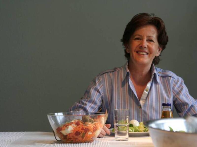 Una mujer sonríe mientras prepara una ensalada saludable en una mesa con una botella de aceite de oliva y un vaso de agua.