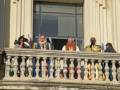 Un grupo de personas, vestidas con trajes coloridos y joyas, saludan desde un balcón de un edificio histórico. La imagen captura una celebración o ceremonia importante, con detalles decorativos y un ambiente festivo.