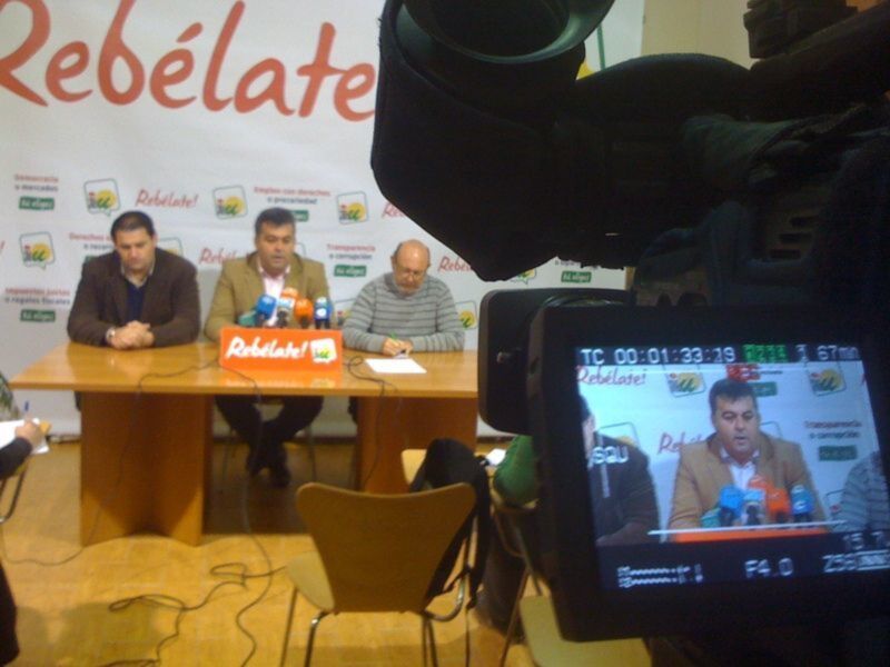 Conferencia de prensa en Rebélate! con tres hombres sentados en una mesa con micrófonos y cámaras de televisión en primer plano.