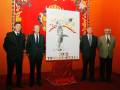 Los hombres están posando frente a un cartel de la feria de toros en Sevilla 2012. El cartel muestra una ilustración con un toro y otros elementos festivos.