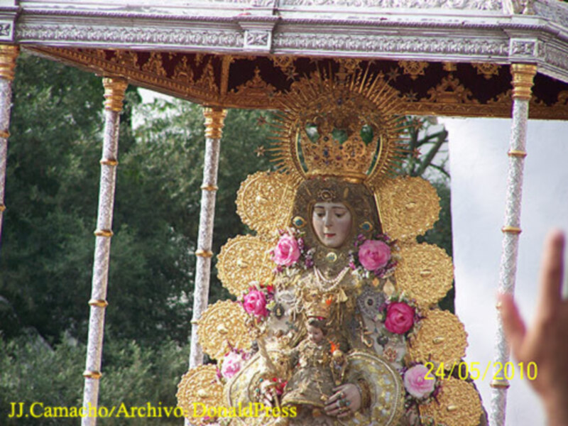 Procesión religiosa con imagen de la Virgen María adornada con flores y joyas, en un día soleado de 2010.