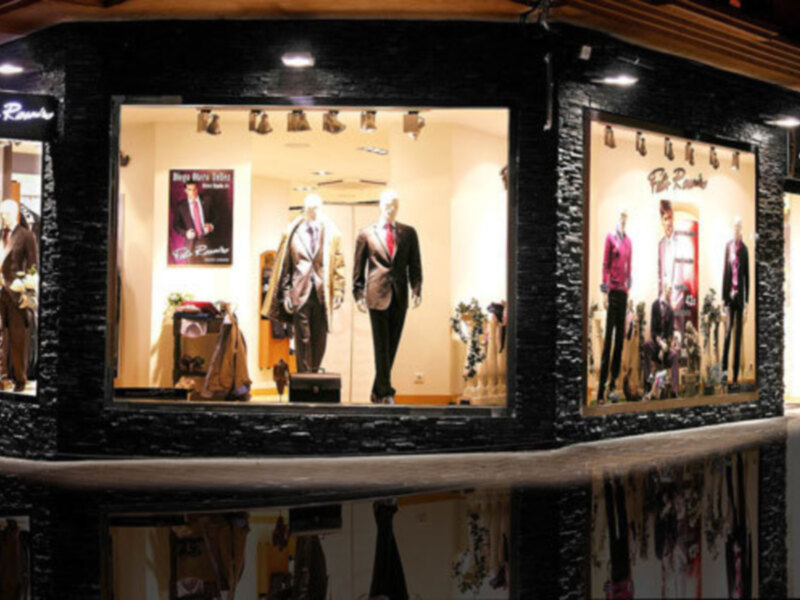 Tienda de ropa con maniquíes vestidos con trajes y chaquetas de lujo, iluminada por luces LED en un escaparate de estilo moderno.