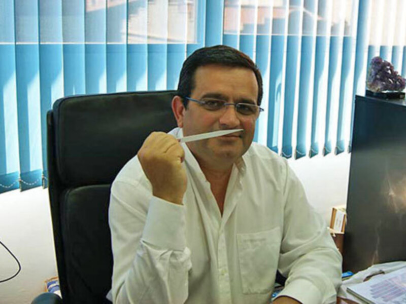 Un hombre en una oficina sosteniendo un cepillo de dientes. La oficina tiene cortinas azules y una computadora en el fondo.