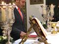 Un hombre en traje negro y corbata roja se inclina sobre una mesa de cristal con velas encendidas. En la mesa, hay un libro abierto en una funda dorada y un candelabro de cristal. En el fondo, se puede ver una pintura enmarcada sobre un muro de color claro.