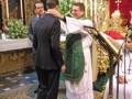 Una ceremonia religiosa en iglesia, con un sacerdote bendiciendo a una persona en traje negro. El altar está decorado con flores y un manto dorado, mientras que una persona en vestido blanco observa la escena.