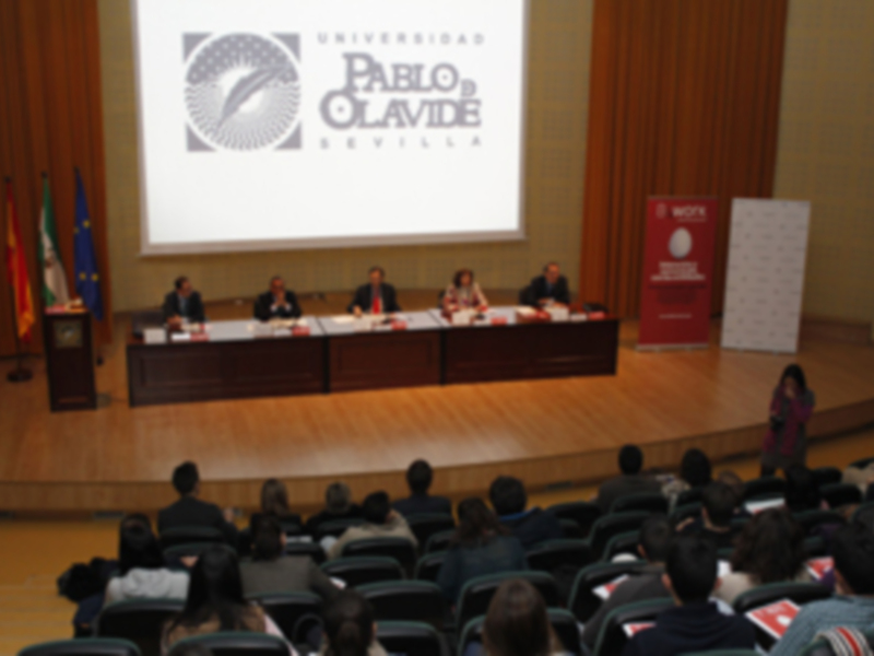 Conferencia en la Universidad Pablo de Olavide, Sevilla, con panelistas discutiendo en el escenario.