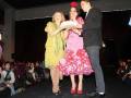 Tres personas en un escenario, dos de ellas vestidas con trajes y falda de flamenco, mientras que la tercera sostiene un bolso. El público observa atentamente desde las butacas.