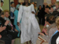 Una mujer vestida con un atuendo blanco y largo camina entre una audiencia de personas que aplauden. La imagen parece ser de un evento formal o ceremonial, posiblemente una boda o ceremonia religiosa.