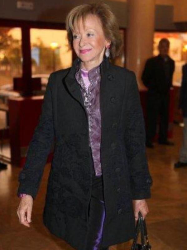 Una mujer camina en un interior bien iluminado, vistiendo un abrigo negro con detalles dorados y una camisa de color lila.