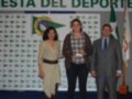 Una mujer en una falda beige sostiene un trofeo mientras está rodeada por dos personas, uno de ellos con una corbata. El fondo muestra un banner del Club Náutico y la bandera de España, junto con el texto "FESTA DEL DEPORTE".