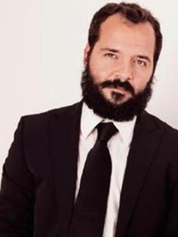 Persona con barba y bigote, vestido con traje negro y corbata, presentando un look formal.