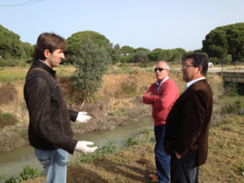 Tres personas discuten cerca de un río en un entorno natural con árboles y vegetación.