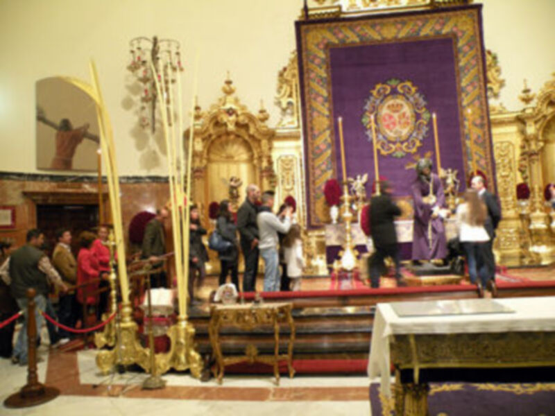 Una iglesia llena de gente, con un altar dorado y una gran tela morada en el fondo. Personas vestidas de rojo y blanco, posiblemente durante una celebración religiosa.