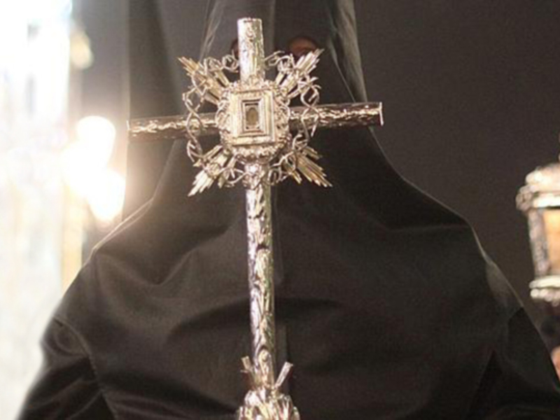 Una cruz de metal con detalles dorados, sostenida por una persona vestida con un hábito negro.