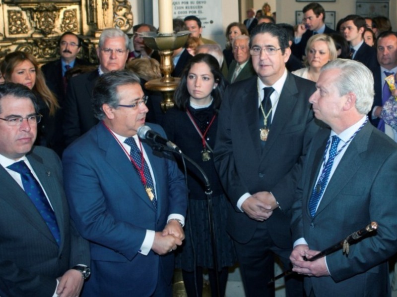Una reunión formal con varios hombres en trajes y mujeres en vestidos, algunos con medallas. El ambiente parece ser un lugar público o institucional, posiblemente una sala de conferencias.