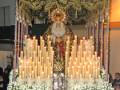 Una estatua de un santo vestido con una saya dorada y un manto negro, rodeado por candelabros de velas encendidas y flores blancas.