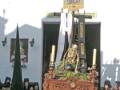 Una procesión religiosa en una iglesia, con un altar central adornado y una imagen de la Virgen en el techo. Personas observan desde afuera, algunas con mantos negros.
